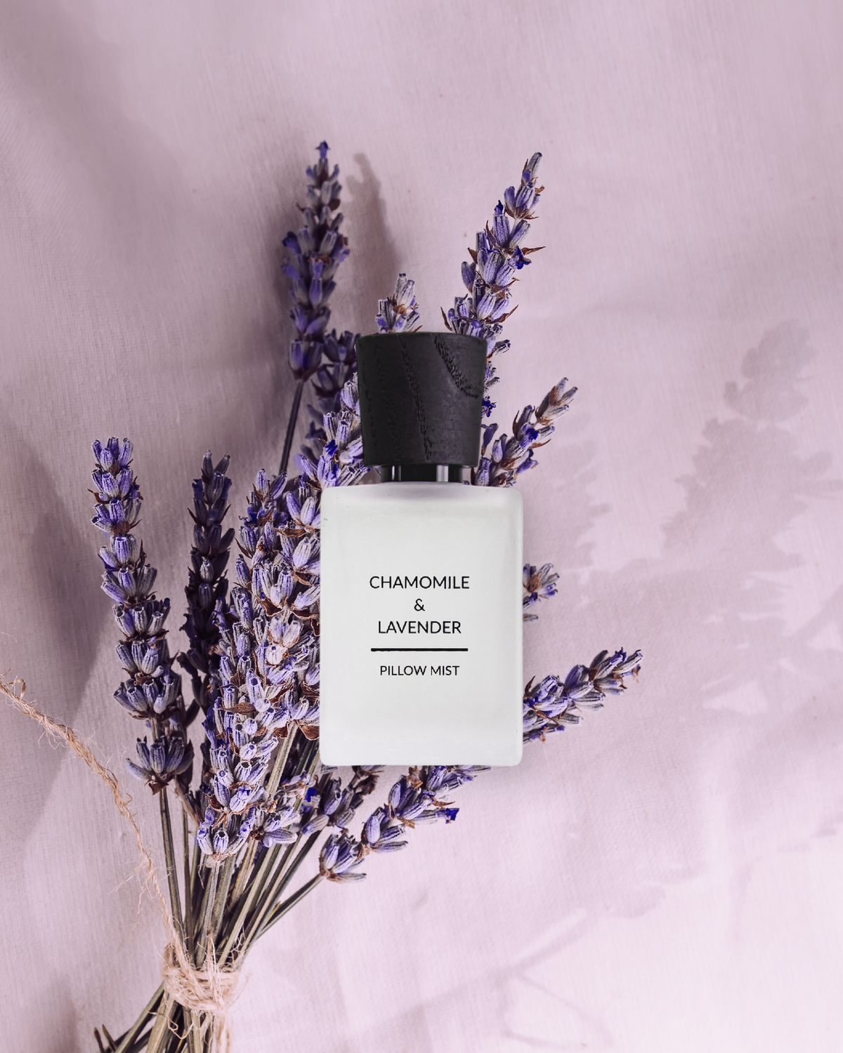 Chamomile and lavender pillow mist bottle surrounded by lavender flowers on a soft pink background